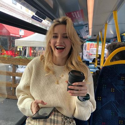 She is smiling at the camera while holding a coffee cup in one hand and her phone in another.